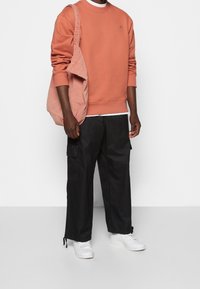 Felpa color ruggine, pantaloni cargo neri, sneakers bianche e una borsa a tracolla rosa chiaro. I pantaloni hanno tasche laterali e lacci regolabili all'orlo.