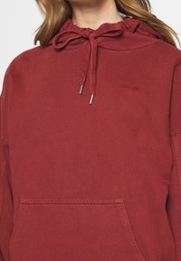Felpa rossa in cotone con cappuccio regolabile, estremità in metallo sui lacci e tasca a marsupio frontale. Logo sottile impresso sul tessuto.