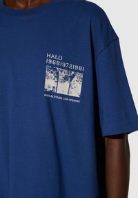 Blå bomullströja med korta ärmar som har en vit grafisk design, inklusive texten "HALO 196819721981" och "HIGH ALTITUDE LOW OPENING."