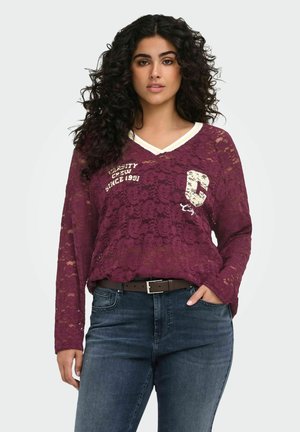 Femme aux cheveux bouclés portant un haut en dentelle bordeaux transparent à manches longues avec un texte et un jean bleu foncé, debout avec une main dans la poche.