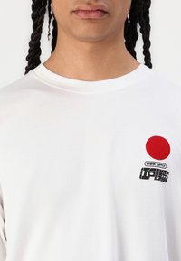 White cotton t-shirt featuring a red circle logo and black graphic text on the left chest. Simple, crew neck design with short sleeves.