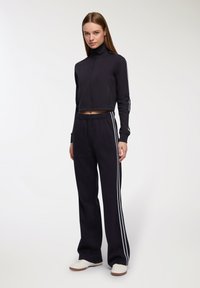 Donna in piedi che indossa una giacca corta nera con zip e pantaloni della tuta neri abbinati con strisce laterali bianche e sneakers bianche.