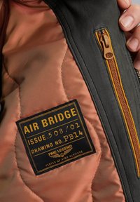 Interno di una giacca di pelle nera con zip dorata; presenta una fodera trapuntata marrone e un'etichetta con la scritta "AIR BRIDGE" e dettagli sul prodotto.