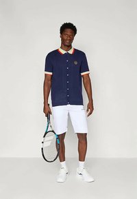 Man in navy blue buttoned polo with red and yellow trim, white shorts, white sneakers, holding a tennis racket against a plain background.