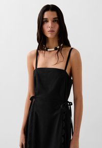 Schwarzes Kleid mit dünnen Trägern, quadratischem Ausschnitt, strukturiertem Stoff, seitlichen Bändern und gewelltem Detail entlang des Saums. Halskette aus weißen und schwarzen Perlen.