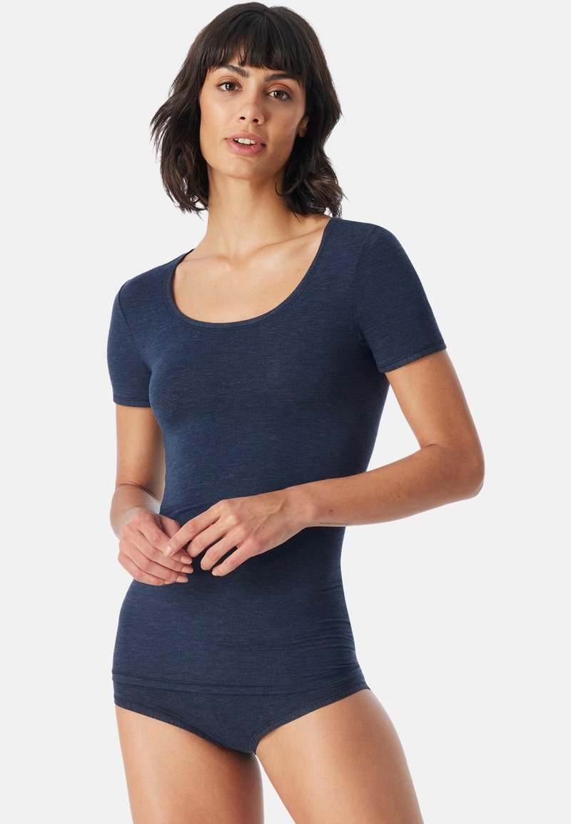 Nainen, jolla on lyhyet tummat hiukset, pukeutuneena istuvaan tummansiniseen lyhythihaiseen paitaan ja vastaaviin alusvaatteisiin, vaaleaa taustaa vasten.