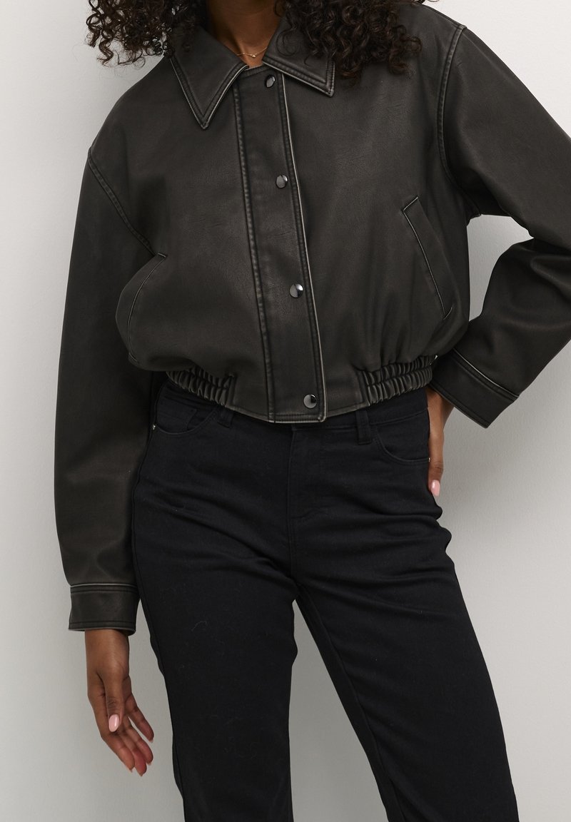Giacca nera in ecopelle tagliata, con ampio colletto, bottoni a scatto e vita elastica. Abbinata a pantaloni neri aderenti.
