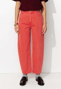 Jean large corail en denim, avec une taille haute, cinq poches et une fermeture à bouton. Portés avec des chaussures sombres et des chaussettes grises.