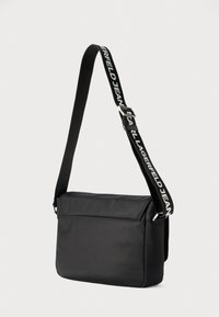 Μαύρη τσάντα ταχυδρόμου από λείο ύφασμα. Διαθέτει ρυθμιζόμενο λουράκι με λογότυπο και κλείσιμο με καπάκι. Απλή ορθογώνια μορφή.