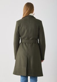 Olajzöld trench coat, övvel a deréknál, strukturált vállakkal és sima gyapjú textúrával. Jellemzője a kivágott gallér és a közepes hosszúságú fazon.