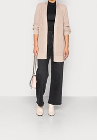 Cardigan beige en tricot à devant ouvert, manches longues, associé à un jean noir à jambes larges et des bottines crème, portant un petit sac à main.
