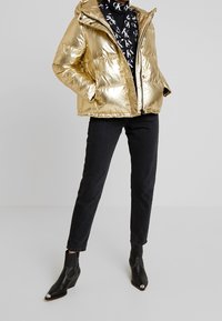 Gouden metallic pufferjack met capuchon, ritssluiting en zijzakken, gecombineerd met zwarte jeans en zwarte laarzen met puntige hak.