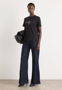T-shirt en coton noir avec le logo "Calvin Klein", associé à un jean large bleu foncé et des escarpins noirs à talons pointus, tenant un sac en cuir noir.