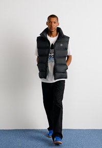 Sort puffervest med forlommer, båret over en hvid T-shirt med et blåt logo, kombineret med sorte bukser og blå sneakers.