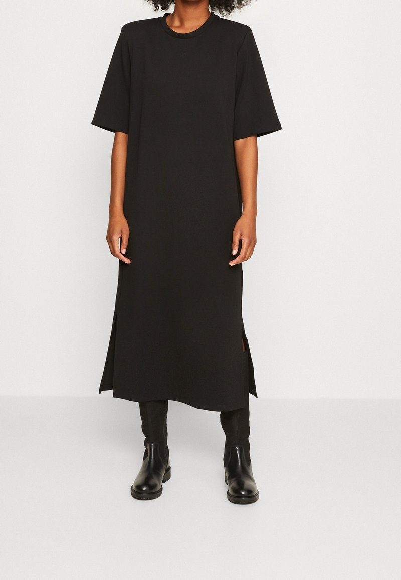 Schwarzes Baumwoll-T-Shirt-Kleid mit kurzen Ärmeln, Rundhalsausschnitt und seitlichen Schlitzen, kombiniert mit schwarzen Knöchelstiefeln, glatte Textur.