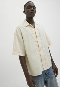 Camisa de manga corta de color beige claro, hecha de una tela suave, que presenta un cierre frontal con botones y un patrón decorativo de ojales a lo largo de la tapeta y los puños.
