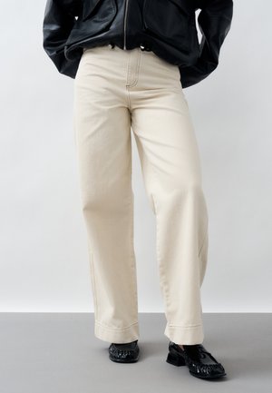 Persona che indossa pantaloni crema a gamba larga, giacca di pelle nera e mocassini neri lucidi, in piedi davanti a uno sfondo semplice.