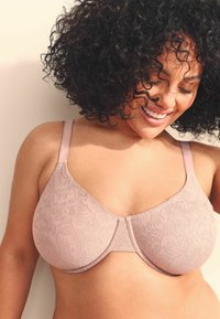 Soutien-gorge en dentelle rose clair avec un design sans coutures, des surpiqûres apparentes, des bretelles réglables et des détails de motifs floraux sur le tissu.