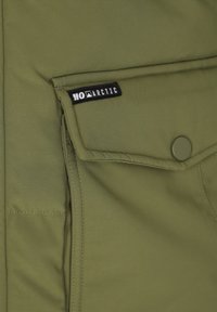 Olivengrøn jakke sektion med en klaplomme, der har en trykknap og et sort mærke med teksten "MOARCTIC." Glat stofoverflade.