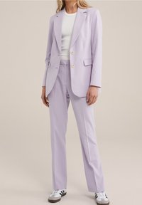 Kvinna iklädd en ljuslila dräkt med skräddarsydd kavaj och byxor, vit topp och vita sneakers med svarta ränder.