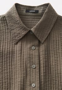 Camicia colletto in tessuto verde oliva testurizzato, con pieghe verticali, sei bottoni e chiusura frontale. Presente etichetta del marchio.