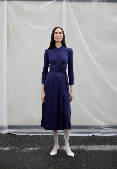 Tamnoplava haljina na preklop s ovratnikom i gumbima, dugih rukava, teksturiranog materijala. Uparena s bijelim čipkastim hulahupkama i bijelim cipelama šiljastih prstiju.
