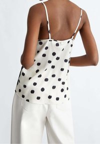 Camisole en satin couleur crème avec des bretelles fines, ornée d'un motif de pois noirs, et dotée d'une fermeture à bouton au dos. Associée à un pantalon blanc.