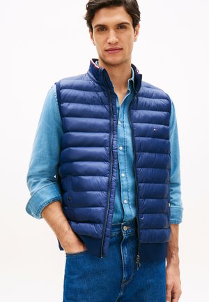 Mann trägt blaue gesteppte Weste über hellblauem Jeanshemd und blauer Jeans, mit einer Hand in der Tasche, stehend vor einfachem weißen Hintergrund.