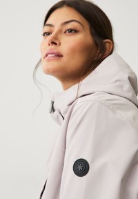 Chaqueta con capucha de color rosa claro y textura suave, con un logo circular en negro en la manga izquierda y cierre de cremallera.