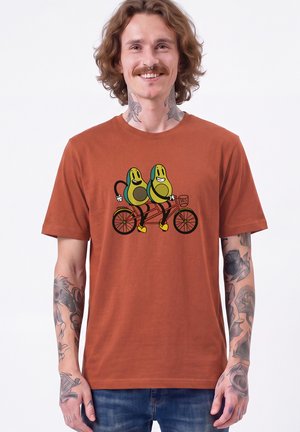 Homme avec des tatouages et un piercing au septum portant un t-shirt couleur rouille avec des avocats dessinés en train de faire du vélo tandem.