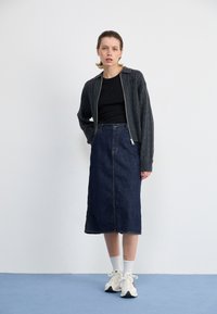 Grijze gebreide ritsjack over een zwarte top, gecombineerd met een lange spijkerrok. Schoeisel: witte sneakers met zwarte accenten. Achtergrond is eenvoudig.
