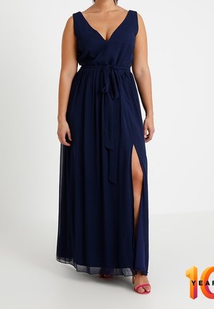 Vrouw draagt mouwloze marineblauwe maxi-jurk met diepe V-hals, taillestrik en hoge dij-split, gecombineerd met roze open-teen hakken.