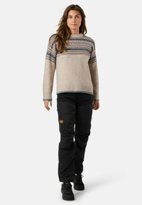 Beiger Strickpullover mit gemustertem Einsatz in Blau und Pink, kombiniert mit schwarzen Hosen mit Taschen und einem Lederlogo-Detail.