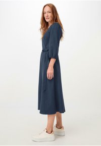 Dunkelblaues, knielanges Kleid mit dreiviertellangen Puffärmel und einem gebundenen Taillendetail. Getragen mit weißen Sneakers, die braune Akzente haben.