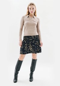 Beige neulepusero pitkillä hihoilla, kauluksella ja nappidetaileilla. Musta kukallinen hame, jossa on kuminauha vyötäröllä, yhdistettynä mustiin polven ylle ulottuviin saappaisiin.