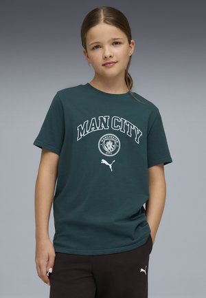 Młoda osoba ubrana w ciemnozieloną koszulkę Manchester City i czarne spodnie Puma, stojąca na tle szarego gradientu.