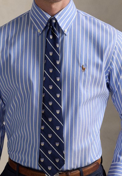 Camisa de riscas azul claro com colarinho abotoadinho, combinada com uma gravata azul-marinho com riscas diagonais e padrões de escudos. Cinto castanho visível.