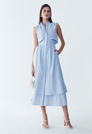 Mujer con vestido midi sin mangas con rayas azules y falda con capas, llevando bolso blanco y sandalias blancas de tacón, de pie sobre fondo blanco.