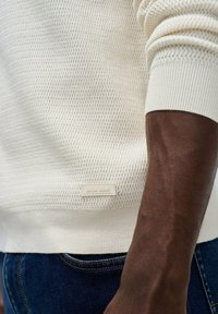 Blizak pogled na teksturiranu kremastu rukavicu i rub svijetle kremaste vune s oznakom "Salsa Jeans", nosi se s tamnoplavi trapericama na tamnoj koži ruke.