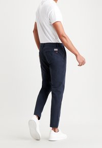 Námořnické chinos kalhoty s tapered střihem, bavlněná tkanina, přední a zadní kapsy, značka na zadní straně, stylizované s bílým tričkem a teniskami.