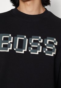 Sudadera negra con texto pixelado "BOSS" en gris y blanco roto, con cuello redondo y puños acanalados. Textura de tela suave.