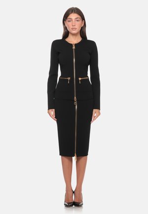 Donna in piedi che indossa un tailleur gonna nero aderente con cerniere dorate sul davanti e sulle tasche, abbinato a décolleté nere.
