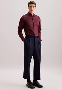 Camicia da uomo a maniche lunghe color bordeaux con colletto a punta, abbinata a pantaloni a pieghe navy a vita alta e scarpe eleganti in pelle nera.