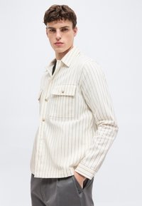 Νέος άνδρας με κοντά καστανά μαλλιά, που φοράει ένα κρεμ πουκάμισο με μακριά μανίκια και κάθετες ρίγες και γκρι παντελόνι, έχοντας το ένα χέρι στην τσέπη.