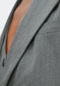 Sacou gri cu textură fină, revere ascuțite și un buzunar la piept. Materialul pare ușor, cu un model subtil de țesătură.