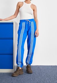 Blå sweatpants med vita vertikala ränder, dragsko i midjan och sidfickor, kombinerat med bruna sportskor och en vit linne.