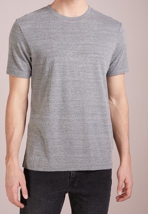 Homme portant un t-shirt gris uni à manches courtes et un jean noir, debout devant un fond neutre.