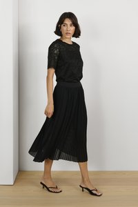 Černý krajkový top s krátkým rukávem, v kombinaci s černou maxi sukní s plisováním. Model nosí černé sandály na podpatku. Jednoduchý, elegantní design.
