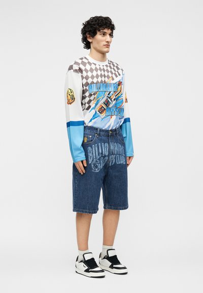 Νεαρός άνδρας που στέκεται, φοράει ένα μακρυμάνικο πουκάμισο με σχέδιο σκακιέρας και αστραπής, τζιν σορτς και μαύρα-άσπρα αθλητικά παπούτσια.