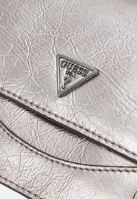 Sølvtekstureret læder med syede sømme og en lille trekantet metallisk Guess USA-logo plade.